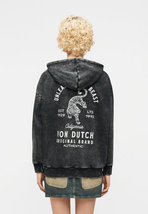 Osoba s kudrnatými blond vlasy nosí černou mikinu s kapucí, na zádech je tygří grafika a nápisy "Unleash Beast" a "Von Dutch Original Brand".