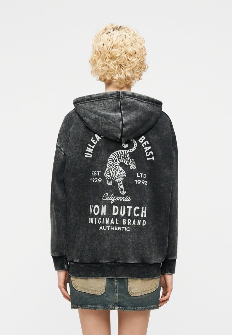 Άτομο με κατσαρά ξανθά μαλλιά που φοράει μαύρο φούτερ με κουκούλα, με γραφικό τίγρης και το κείμενο «Ξεσπάστε το θηρίο» και «Von Dutch Original Brand» στην πλάτη.