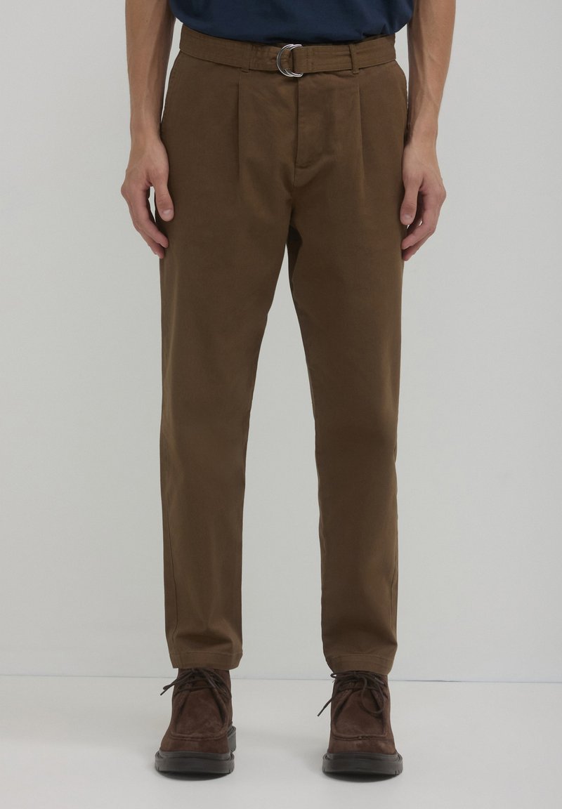 Pantaloni a pieghe marroni con vita aderente e fibbia argentata, abbinati a scarpe marroni stringate, con una texture liscia.