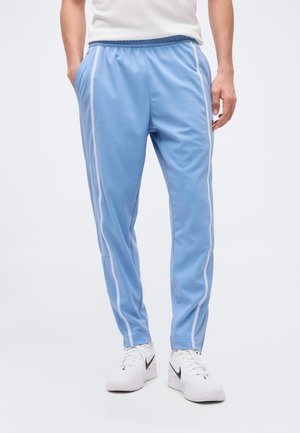 Muž v svetlomodrých športových nohaviciach s bielymi bočnými pruhmi a bielych teniskách Nike, s rukami vo vreckách, stojaci na hladkom pozadí.