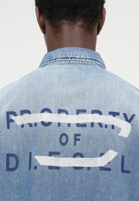 Ανοιχτό μπλε τζιν μπουφάν με μεγάλο, σκούρο μπλε κείμενο στο πίσω μέρος που γράφει "PROPERTY OF D.I.E.S.E.L", με λευκές λεπτομέρειες πάνω από το κείμενο.