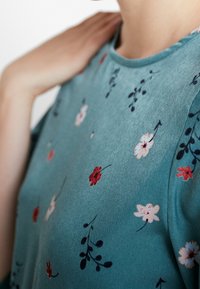 Turkos kortärmad topp med en broderad blommönster i rött och rosa med gröna stjälkar, tillverkad av ett slätt tyg med en subtil lyster.