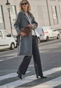 Grijze wollen jas met een getailleerd ontwerp, licht overhemd eronder, donkere broek met wijde pijpen en zwarte laarzen met hoge hakken. Bruine schoudertas.