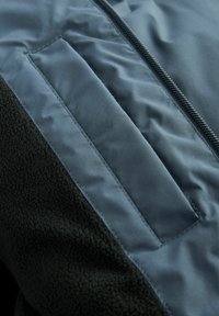 Detail fleecové a nylonové bundy ukazující tmavě modrou zipovou část a texturovanou černou fleecovou panel s plochým designem kapsy.