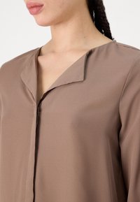 Blouse en satin légère et brun avec un décolleté en V et un éclat subtil. Présente une simple patte de boutonnage et une texture lisse.