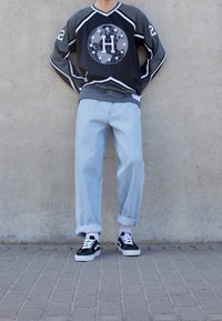 Person in einem schwarz-grauen Langarmshirt mit einem Globus-Emblem, hellblauen hochgekrempelten Jeans und schwarzen Turnschuhen, der an einer Wand steht.