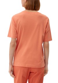 Person med kort brunt hår iført en ensfarvet orange kortærmet t-shirt og matchende orange bukser set bagfra.