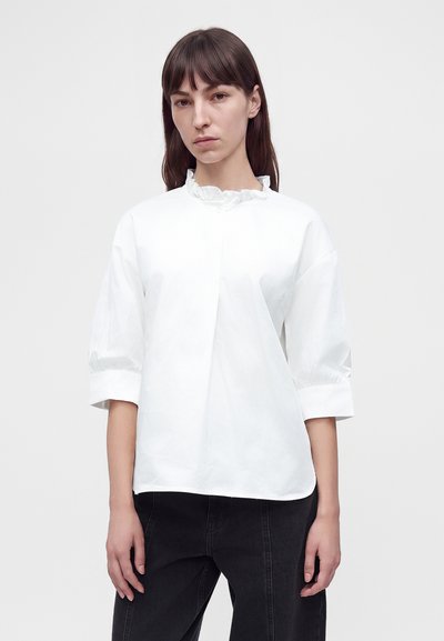 Femme aux cheveux bruns et raides portant un chemisier blanc à col volanté et manches trois-quarts ainsi qu’un pantalon noir sur un fond uni.