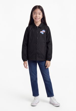 Lachend meisje met lang donker haar, gekleed in een zwart jasje met knopen en logo, een blauwe jeans en witte hoge sneakers, staand tegen een witte achtergrond.