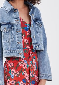Veste en jean délavée claire, au design court, avec des boutons argentés, associée à une combinaison short rouge à motifs floraux roses et bleus.