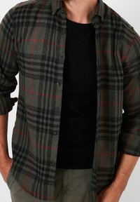 Man som bär en mörkgrön och svart rutmönstrad skjorta med knappar över en svart t-shirt med uppvikta ärmar och olivgröna byxor.