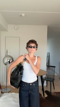 Bolso de mano de cuero negro, de estilo holgado, con un borde recogido y acentos de cadena dorada. El atuendo casual incluye una camiseta sin mangas blanca y unos jeans de mezclilla oscuros.