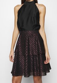 Svart halterneck klänning med en skiktad kjol, som har ett zickzackmönster i svarta och rosa paljetter. Satinfinish med en åtsittande bodice.