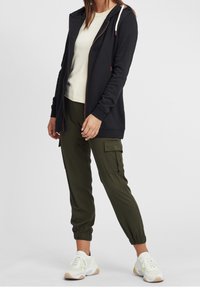 Felpa con zip blu scuro e cappuccio con cordino, abbinata a una maglietta beige chiaro e pantaloni cargo verde oliva, completata da sneakers bianche.
