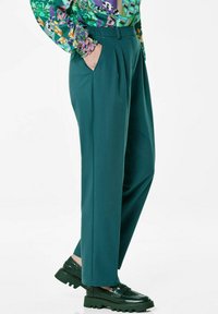 Turquoise maatbroek met een hoge taille, plooien en zijzakken. Gecombineerd met zwarte chunky loafers voor contrast. Gladde stoftextuur.