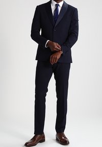 Marinblå kostym med kavajslag i siden och spetsig form, smal passform, matchande mörk slips och bruna läderskor. Skräddarsydda byxor kompletterar utseendet.