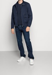 Marinblå zip-jacka med ståkrage, två framfickor, kombinerad med mörkblå jeans och vita sneakers på en neutral bakgrund.