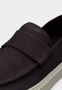 Σκούρο καφέ δερμάτινο παπούτσι τύπου slip-on με φαρδιά λεπτομέρεια από λουρί και λευκή λαστιχένια σόλα, εμφανίζεται σε κοντινή λήψη.