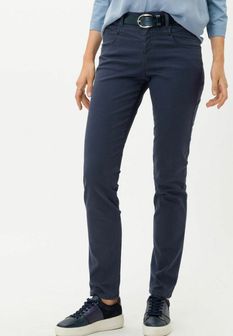 Pantalons slim-fit bleu marine foncé en mélange de coton doux, avec des poches avant et une ceinture avec boucle en argent. Associés à des baskets sombres.
