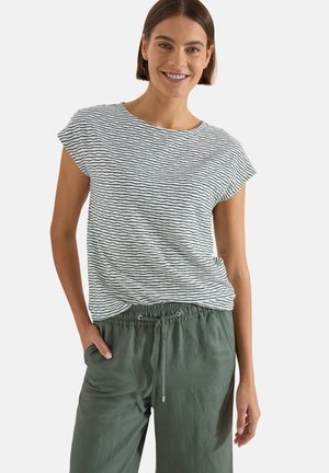 Femme souriante aux cheveux bruns courts portant un t-shirt blanc à rayures bleu marine à manches courtes et un pantalon vert olive avec cordon.