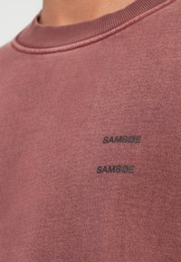 Sudadera burdeos con una textura sutil, que presenta un cuello acanalado y dos etiquetas impresas "SAMSOE" en negro en la parte delantera.