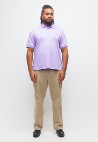 Άνδρας με ντρέντλοκς φοράει ανοιχτό μωβ πόλο πουκάμισο, μπεζ παντελόνι και μαύρα παπούτσια, στέκεται μπροστά σε λευκό φόντο.