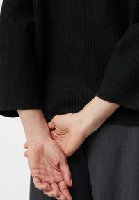 Czarny prążkowany sweter z szerokimi rękawami, noszony z szarymi spodniami. Ręce modelki złożone za plecami, ukazujące teksturę i kształt skóry.