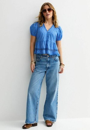 Vrouw draagt een blauwe blouse met pofmouwen, jeans met wijde pijpen, oversized zonnebril en bruine sandalen met open tenen, met armbanden aan beide polsen.