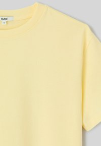 T-shirt en coton jaune pâle à manches courtes avec encolure arrondie, taille petite, étiquette de la marque BZB visible à l'intérieur du col.
