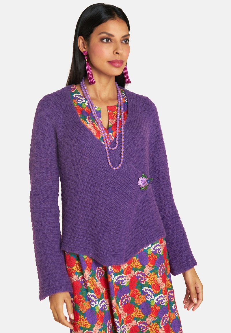 Purple knit sweater with a v-neck, textured fabric, and long sleeves. Features a green flower accent. Paired with a colorful floral dress.