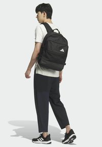 Schwarzer Rucksack aus Stoff mit mehreren Reißverschlussfächern, verstellbaren Trägern und einem weißen Logo auf der Vorderseite.