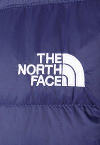 Logo "THE NORTH FACE" blanc brodé sur un tissu matelassé bleu foncé, probablement partie d'une veste ou d'un vêtement de plein air.