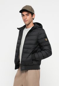 Young man wearing checked cap, black puffer jacket with hood, light gray shirt, and beige pants, standing with hands in pockets.