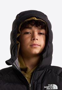 Jeune personne aux cheveux bouclés portant une veste noire à capuche par-dessus un sweat à capuche vert olive, face à nous avec une expression neutre.