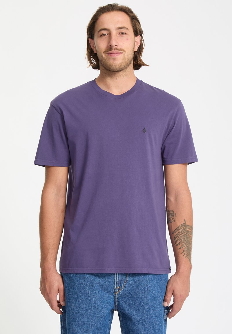 Lila Baumwoll-T-Shirt mit kurzen Ärmeln und rundem Ausschnitt. Verfügt über ein kleines schwarzes Logo auf der Brust. Getragen mit blauen Jeans.