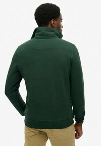 Sudadera de forro polar verde oscuro con cuello alto, puños ribeteados y dobladillo. Parece suave con una textura lisa, diseñada para comodidad y calidez.