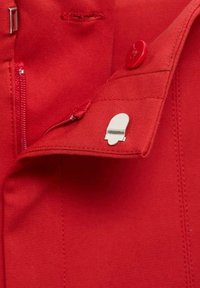 Červená tkanina s hladkým povrchom obsahuje striebornú odznak, červený gombík a zipsový detail na golieri.