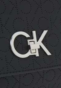 Sort taske i kunstlæder med prægede cirkulære mønstre, en sølvfarvet CK-logo hardware detalje og syninger langs kanterne.