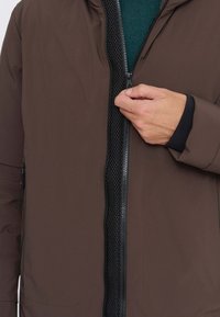 Braune Jacke mit glatter Außenseite, schwarzem Reißverschluss vorne, Mesh-Futter und Vorderentaschen. Verfügt über eine Kapuze und elastische Bündchen für eine gute Passform.