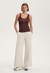 Rjava rebrasta majica na nit s krojem z zaobljenim izrezom in tremi gumbnimi detajli, kombinirana z ohlapnimi, kremnimi širokimi hlačami in vsakodnevnimi športnimi copati.