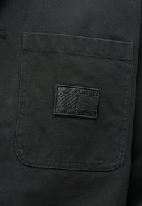 Μαύρο ύφασμα με υφή διαθέτει τσέπη στο αριστερό μέρος του στήθους και ραμμένη ορθογώνια επιφάνεια που παρουσιάζει ένα γεωμετρικό σχέδιο.