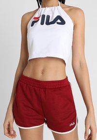 Mujer vestida con un top corto halter blanco de FILA y pantalones cortos deportivos rojos de FILA con ribete blanco, posando con los brazos relajados a los lados.