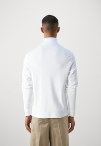 Chemise blanche à manches longues avec un col haut, texture lisse et coupe ajustée, associée à un pantalon beige, vue de l'arrière.