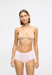 Vrouw met kort bruin haar, gekleed in een nude beha en lichtroze high-waisted onderbroek met kantafwerking, staande tegen een simpele witte achtergrond.