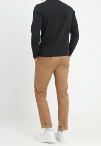 Mężczyzna w czarnej koszuli z długimi rękawami, w beżowych dopasowanych, podwiniętych spodniach, w białych, wysokich trampkach, stoi z rękami w kieszeniach, odwrócony plecami.