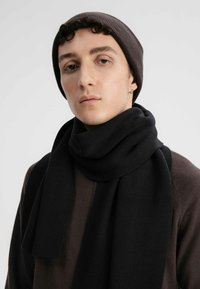 Zwarte gebreide sjaal om de nek gewikkeld, gecombineerd met een bruine geribbelde beanie. De sjaal heeft een zachte, dikke textuur en een eenvoudig ontwerp.