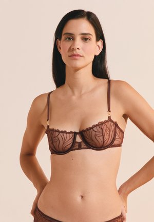 Donna con capelli scuri che indossa un reggiseno in pizzo marrone trasparente con motivi floreali su uno sfondo chiaro uniforme.