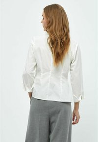 Blusa bianca con maniche lunghe, design aderente, caratterizzata da polsini piegati e una texture liscia. Tessuto semplice, senza motivi o dettagli visibili.