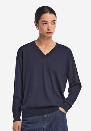 Kvinde iført en marineblå V-hals sweater og blå jeans står med den ene hånd i lommen og kigger til siden mod en ensfarvet baggrund.