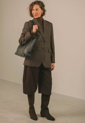 Donna che indossa un blazer marrone a quadri, pantaloni a zampa d'elefante abbinati, stivali scuri e una borsa nera in pelle portata sulla spalla.